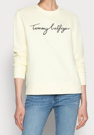 Person wearing a pale yellow sweatshirt with "Tommy Hilfiger" script and light blue jeans, standing with hands relaxed by sides.