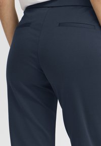 Sötétkék nadrág sima anyagból, két hátsó zsebbel, lapos kialakítással. A derékrész egyszerű és szabott.