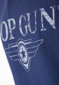 Marinblå bomullströja med vit sliten text "TOP GUN" och ett emblem med vingar och en stjärna, som visar en avslappnad design.