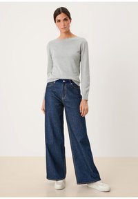 Lichtgrijze gebreide trui met een brede halslijn, gecombineerd met donkerblauwe wijde jeans en witte sneakers, tegen een neutrale achtergrond.
