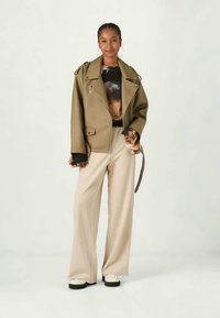 Oliwkowa, oversize kurtka z klapami i zamkiem, czarna bluzka we wzory pod spodem, beżowe spodnie szerokie w nogawkach oraz białe buty.