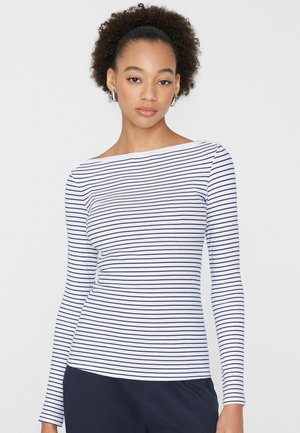 Frau mit lockigem Haar, die silberne Creolenohrringe und ein weißes Langarmshirt mit dünnen horizontalen dunkelblauen Streifen trägt, steht vor einem weißen Hintergrund.