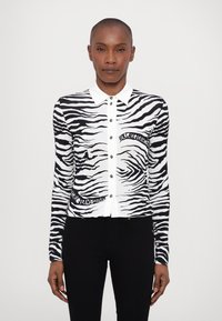 Juostelėta zebra rašto džemperis juodai baltos spalvos, su sagomis priekyje, apykakle ir dekoratyviniu tekstu apatiniame kairiajame kampe.