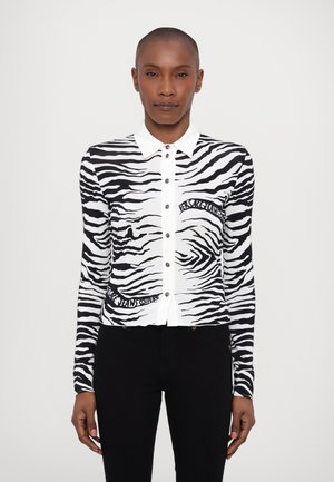 Zebra rakstu kardigāns melnbaltā krāsā ar pogu aizdari, apdari ap kaklu un dekoratīvu teksta detaļu apakšējā kreisajā pusē.
