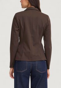 Temno rjav prilagojen blazer z jasno obliko, dolgimi rokavi in rahlim sijajem. Nošen preko modrega jeansa, ki poudarja ukrivljene šive in prilegajoč kroj.