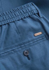 Pantalon en tissu bleu avec une ceinture élastique, une poche arrière boutonnée et une petite étiquette métallique portant l'inscription "QUINTESSENCE."