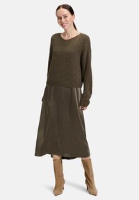 Frau in olivgrünem Strickpullover über schimmerndem olivgrünem Midirock mit beigen Wildleder-Stiefeletten, stehend und leicht zur Seite lächelnd.