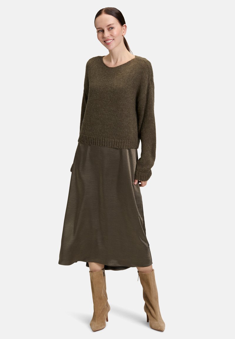 Frau in olivgrünem Strickpullover über schimmerndem olivgrünem Midirock mit beigen Wildleder-Stiefeletten, stehend und leicht zur Seite lächelnd.