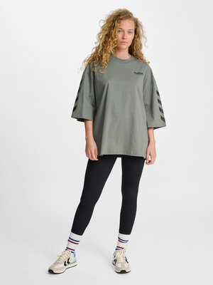 Jonge vrouw met krullend haar die een oversized groen t-shirt, zwarte legging, gestreepte sokken en beige sneakers draagt, staand tegen een effen achtergrond.