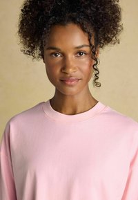 Svetlo roza sweatshirt iz mehkega blaga, z okroglo izreznico in sproščenim krojem. Model ima kodraste lase. Enostavna zasnova, brez vzorcev ali detajlov.