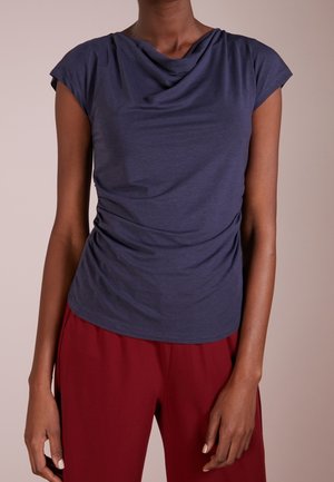 Person trägt ein marineblaues kurzärmeliges drapiertes Oberteil und rote Hose, Arme locker seitlich am Körper vor neutralem Hintergrund.