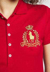 Chemise polo rouge en tissu texturé. Présente un logo brodé doré avec un motif de cheval et de laurier, et une fermeture à boutons blancs.