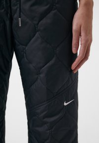 Černé prošívané kalhoty s lesklým povrchem, sází na stříbrné logo Nike na spodní straně a šňůrku v pase.