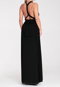Femme portant une longue robe noire dos nu avec des bretelles croisées et un nœud noué dans le bas du dos, debout devant un fond uni.