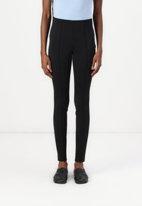 Leggings noirs taille haute avec un ajustement serré, présentant des coutures avant et un tissu lisse et extensible. Portés avec des mocassins noirs.