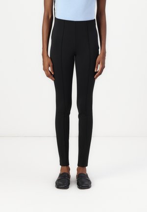 Leggings noirs taille haute avec un ajustement serré, présentant des coutures avant et un tissu lisse et extensible. Portés avec des mocassins noirs.