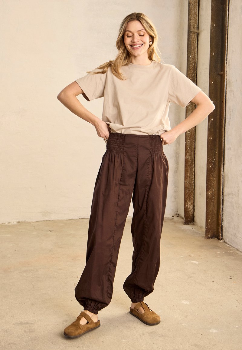 Femme souriante ajustant un pantalon marron foncé à taille élastique, portant un t-shirt beige et des chaussures marron sans lacets, debout sur un sol en béton près d'un mur.