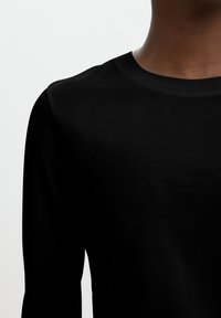 Persona con una maglietta nera semplice a maniche lunghe e scollo rotondo su uno sfondo chiaro uniforme.