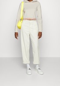 Kobieta w kremowej bluzie z kapturem, białych spodniat z mankietami, białych trampkach Calvin Klein, białych skarpetach i nosząca jasnółtą torbę na ramię.