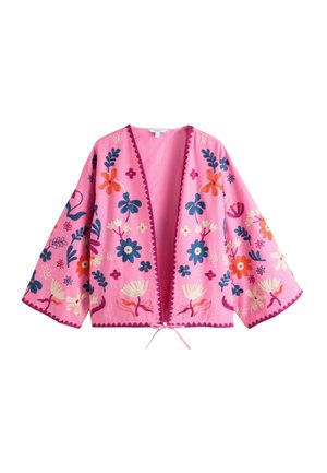 Kvinde med langt krøllet hår står på en strand iført cremefarvede bukser og et pink blomsteret sjal under en klar blå himmel.