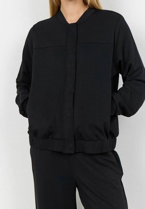 Femme portant une veste bomber noire zippée et un pantalon noir assorti, debout les mains dans les poches de la veste devant un fond uni.
