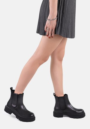 Stivali neri alla caviglia realizzati in pelle liscia, con pannelli elasticizzati laterali e suole in gomma robuste, abbinati a una gonna grigia plissettata.