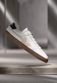 Adidași din piele albă cu un accent bej, călcâi bleumarin și talpă din cauciuc maro. Prezintă detalii cusute și un logo distinctiv.