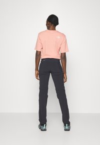 T-shirt en coton rose avec logo dans le dos, associé à un pantalon stretch noir. Le modèle porte des chaussures de randonnée grises et turquoise. Design simple et décontracté.