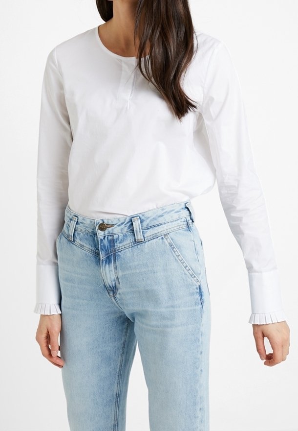 Chemise blanche à manches longues avec un col rond, dotée de poignets plissés. Associée à un jean en denim bleu clair à taille haute et coupe décontractée.