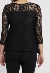 Haut en dentelle noire à manches longues, présentant des motifs floraux élaborés et une coupe décontractée, associé à un pantalon noir côtelé.