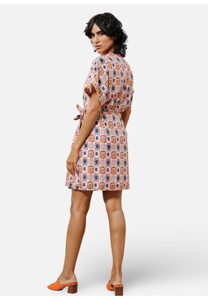 Korte mouw, bedrukte jurk in pastelkleuren met een strikceintuur. Bevat geometrische vormen en bloemaccenten. Gegdraag met oranje schoenen.