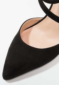 Chaussure en daim noir à bout pointu avec bride à la cheville portée sur un pied à teint clair, présentée sur fond blanc.