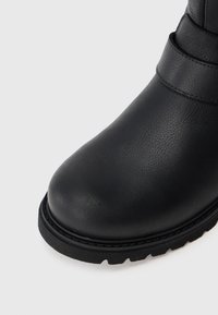 Bota de couro preta com biqueira arredondada, detalhe de banda costurada e sola espessa e robusta sobre um fundo claro e liso.