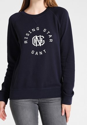 Frau trägt einen marineblauen Sweatshirt mit langen Ärmeln und dem Text "RISING STAR GANT" sowie einem weißen Logo auf der Vorderseite, kombiniert mit grauen Jeans.