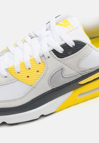 Zapatillas blancas y grises con acentos amarillos, que presentan paneles texturizados, un frente de cordones y una amortiguación visible en suela negra.