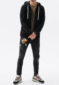 Homme portant un sweat à capuche noir zippé, un t-shirt vert olive, un jean noir déchiré avec des patchs, et des baskets beige, noir et marron, debout les mains jointes.