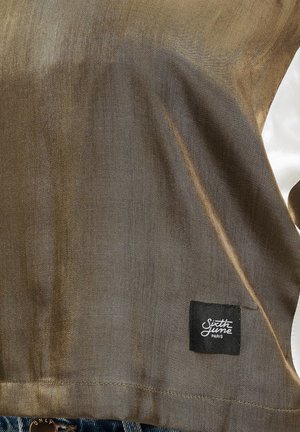 Kortærmet skjorte i skinnende bronze stof med hvide detaljer. Har et sort mærke med "Sixth June Paris" i nederste venstre hjørne.