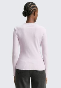 Vrouw met een gevlochten haarbun, gekleed in een lichtroze ribbeltrui met lange mouwen en donkere jeans, van achter bekeken tegen een neutrale achtergrond.