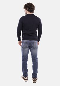 Uomo con capelli ricci che indossa un maglione nero, jeans blu e scarpe da ginnastica Nike marroni, in piedi di spalle contro uno sfondo semplice.