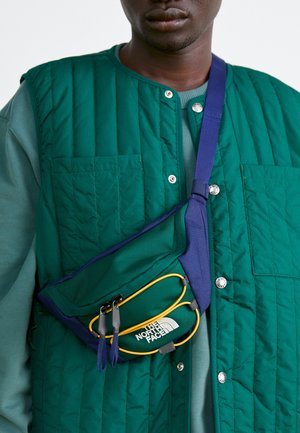 Mężczyzna noszący turkusową pikowaną kamizelkę i torbę na ramię z niebieskimi paskami i żółtymi akcentami, z logo The North Face.