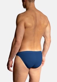 Mannelijke model van achteren afgebeeld in donkere blauwe zwemboxers, staande tegen een effen lichte achtergrond.