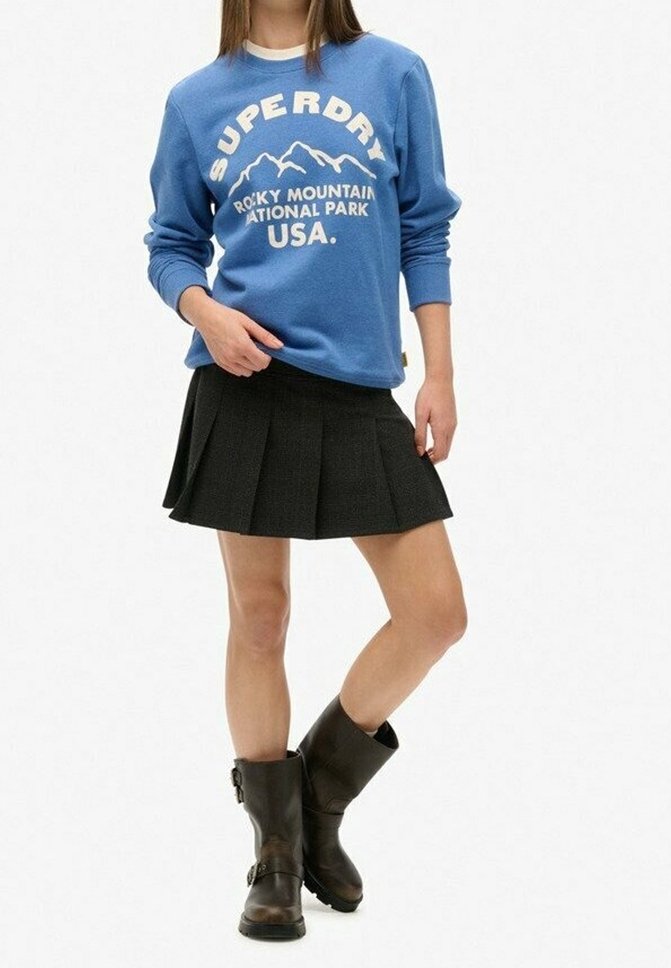 Sudadera azul con el texto blanco "Superdry" y un gráfico de montaña, combinada con una falda negra con pliegues. Botas marrones hasta la mitad de la pantorrilla con detalle de hebilla.
