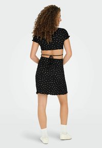 Femme aux cheveux bouclés portant un ensemble deux pièces à pois noirs avec des liens dans le dos, des chaussettes blanches et des baskets blanches, se tenant devant un fond uni.