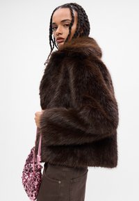 Casaco de pelagem falsa castanho com um colarinho largo, apresentando um tecido texturizado e um corte curto e quadrado. Mala rosa com lantejoulas segurada com uma mão.