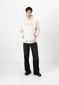 Crème hoodie met het "TOMMY JEANS" logo, voorzien van een kanga-roezak en trekkoorden. Gecombineerd met brede zwarte jeans en zwarte schoenen.