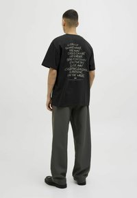 Black cotton t-shirt with oversized fit, featuring a handwritten text design on the back. Paired with loose, dark gray trousers and black shoes.
