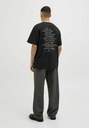 Black cotton t-shirt with oversized fit, featuring a handwritten text design on the back. Paired with loose, dark gray trousers and black shoes.
