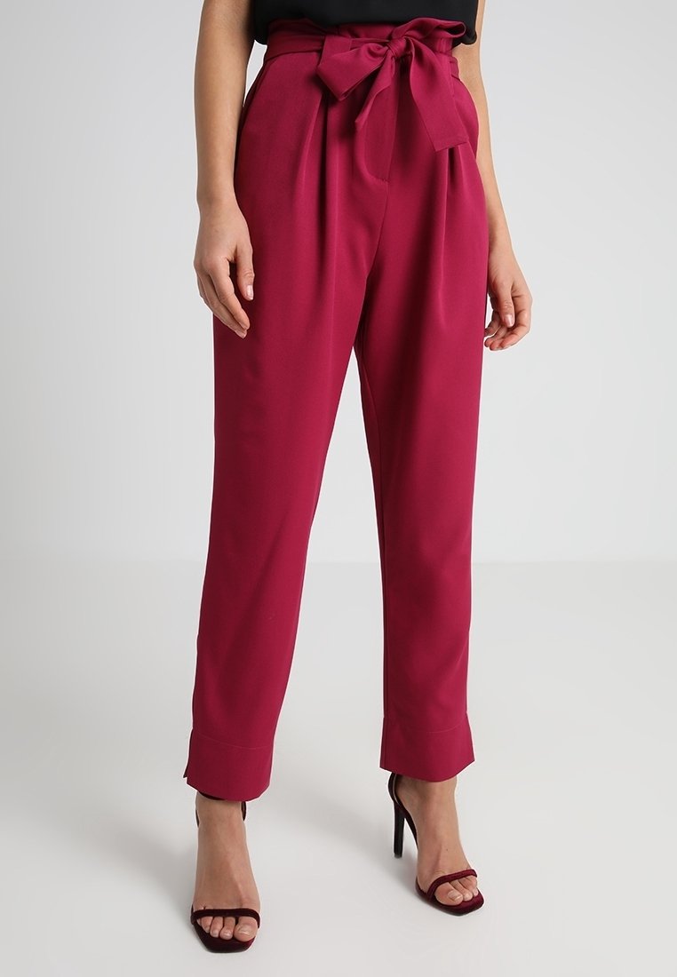 Femme portant un pantalon taille haute magenta avec des plis, une ceinture nouée à la taille, des ourlets retroussés et des sandales à talons hauts assorties.