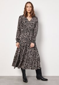 Langes, schwarz-creamfarbenes, gemustertes Kleid mit V-Ausschnitt, langen Ärmeln und gesmokter Taille. Getragen mit schwarzen Cowboys Stiefeln.