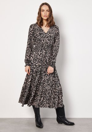 Langes, schwarz-creamfarbenes, gemustertes Kleid mit V-Ausschnitt, langen Ärmeln und gesmokter Taille. Getragen mit schwarzen Cowboys Stiefeln.